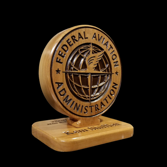 A double-sided desk carving of the Federal Aviation Administration (FAA) logo crafted from cherry hardwood, with the FAA emblem and 'FEDERAL AVIATION ADMINISTRATION' text on one side, and a biplane with 'U.S.